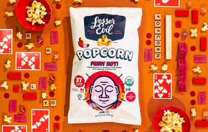 A bag of LesserEvil Fiery Hot Organic Popcorn sits on an orange surface surrounded by popcorn, game cards, Scrabble tiles, tickets, and small bowls.