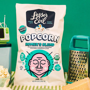A bag of OrganicPopcorn Homers Blend Organic Popcorn sits on a table with popcorn, cheese, a grater, a plant, a mini TV, and a green background.