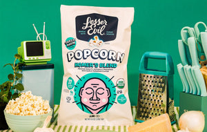A bag of OrganicPopcorn Homers Blend Organic Popcorn sits on a table with popcorn, cheese, a grater, a plant, a mini TV, and a green background.