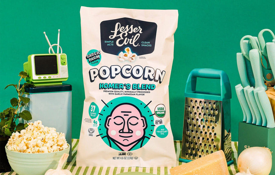 A bag of OrganicPopcorn Homers Blend Organic Popcorn sits on a table with popcorn, cheese, a grater, a plant, a mini TV, and a green background.