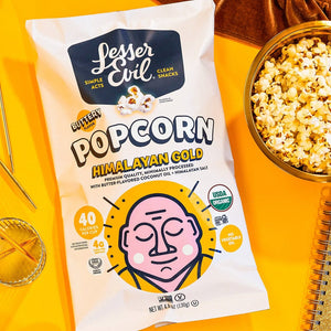 A bag of LesserEvil Himalayan Gold Organic Butter Flavor Popcorn sits on a yellow surface next to a bowl of popcorn, a glass, a gold notebook, and gold paper clips.