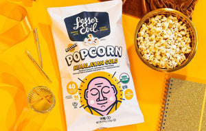 A bag of LesserEvil Himalayan Gold Organic Butter Flavor Popcorn sits on a yellow surface next to a bowl of popcorn, a glass, a gold notebook, and gold paper clips.