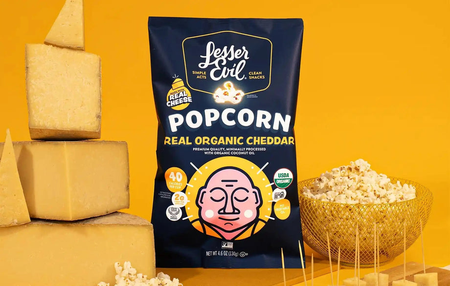 A Big Flavor Popcorn Box by LesserEvil Snacks is surrounded by popcorn, cheddar cheese blocks, and bowls on a yellow background.