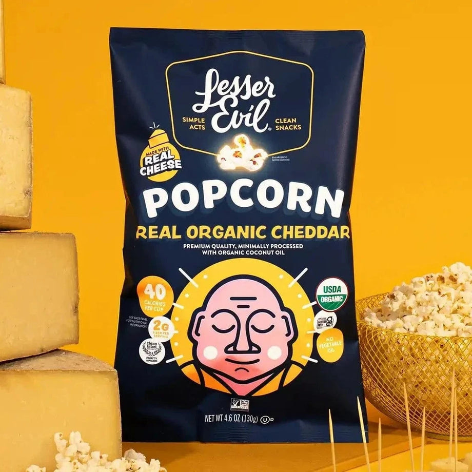A box of LesserEvil Snacks Big Flavor Popcorn stands upright with popcorn and cheese blocks nearby, set against a bright yellow background.