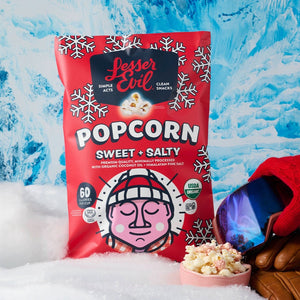 A bag of LesserEvil Sweet + Salty Popcorn, made with organic cane sugar, sits in snow by a bowl of popcorn, ski gear, and mittens on an icy blue background.