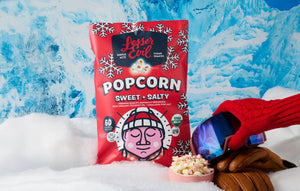 A bag of LesserEvil Sweet + Salty Popcorn, made with organic cane sugar, sits in snow by a bowl of popcorn, ski gear, and mittens on an icy blue background.