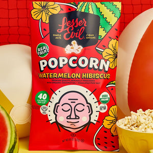 A vibrant bag of LesserEvil Watermelon Hibiscus Popcorn pops among beach balls and watermelon slices, reflecting summer fun and fresh flavors on a bold yellow and red backdrop.