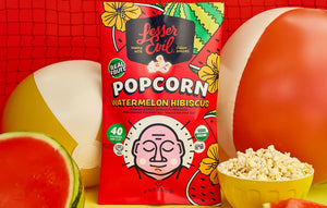 A vibrant bag of LesserEvil Watermelon Hibiscus Popcorn pops among beach balls and watermelon slices, reflecting summer fun and fresh flavors on a bold yellow and red backdrop.
