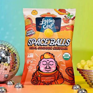 A bag of SpaceBalls Real Organic Cheddar Space Balls is placed between a disco ball and a bowl of organic cheese treats on a colorful gradient background.