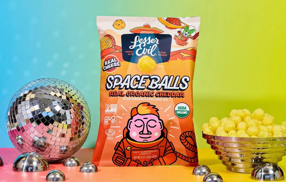 A bag of SpaceBalls Real Organic Cheddar Space Balls is placed between a disco ball and a bowl of organic cheese treats on a colorful gradient background.