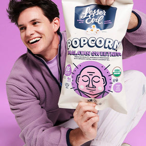 A smiling man in a purple jacket holds a large bag of OrganicPopcorns Himalayan Sweetness Gourmet Popcorn against a vibrant purple background.