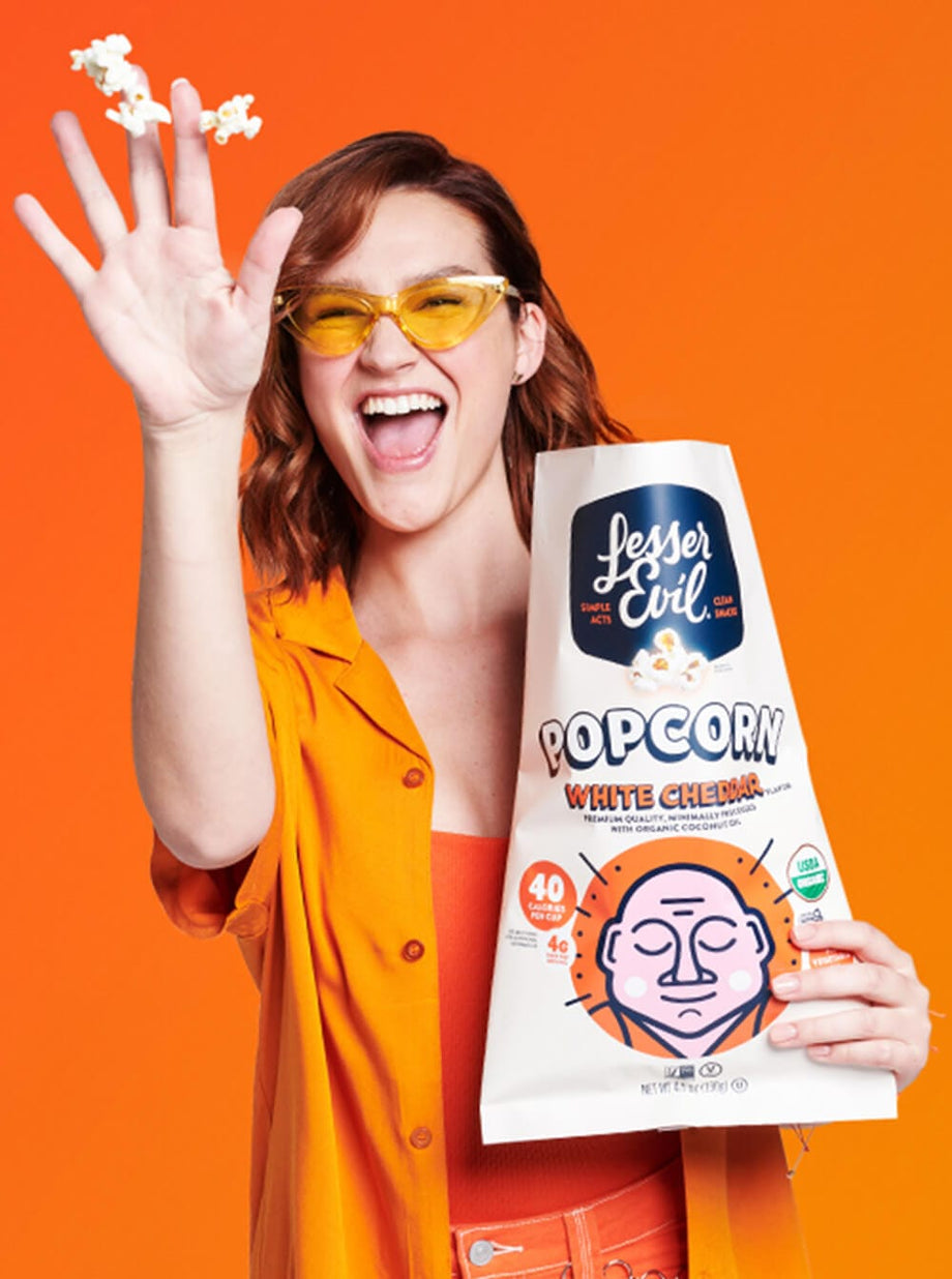 A smiling woman in yellow sunglasses and an orange shirt tosses OrganicPopcorn Organic White Cheddar Flavor Popcorn against an orange background.