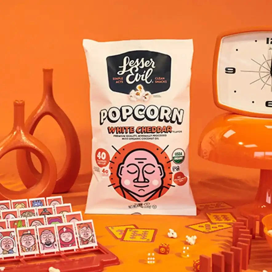 A box of The Classics Popcorn Box by LesserEvil Snacks sits on an orange table, surrounded by popcorn, a retro clock, orange decor, and a matching game with illustrated cards.