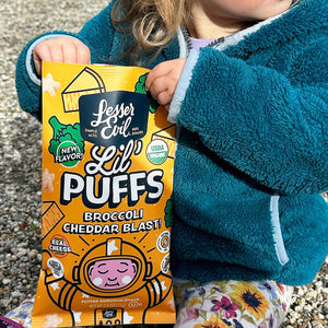 A young child in a teal jacket sits on gravel outdoors, holding a bag of LesserEvil Broccoli Cheddar Blast Lil Puffs snacks.