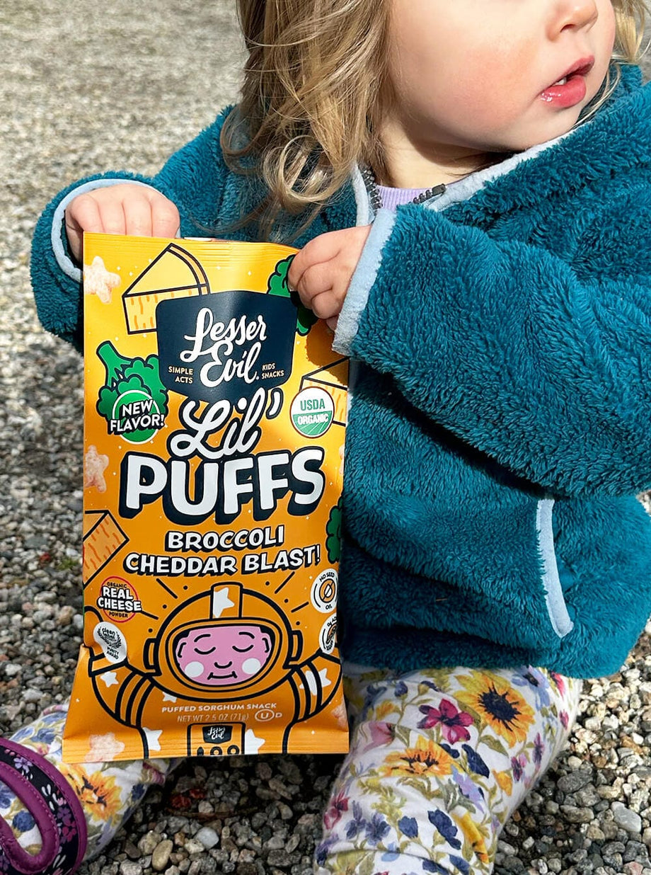 A young child in a teal jacket sits on gravel outdoors, holding a bag of LesserEvil Broccoli Cheddar Blast Lil Puffs snacks.