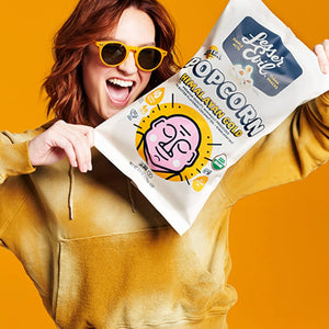 A smiling person in yellow sunglasses and a hoodie holds up a bag of LesserEvil Himalayan Gold Organic Butter Flavor Popcorn against a bright yellow background.
