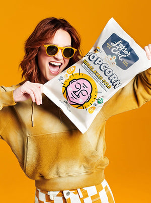 A smiling person in yellow sunglasses and a hoodie holds up a bag of LesserEvil Himalayan Gold Organic Butter Flavor Popcorn against a bright yellow background.