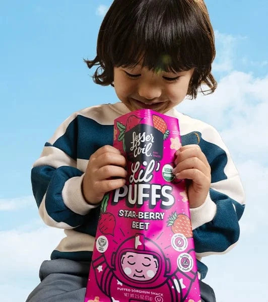 A young child in a striped sweater smiles while holding a large, colorful bag of Lil Puffs Star-Berry Beet snacks against a blue sky background.
