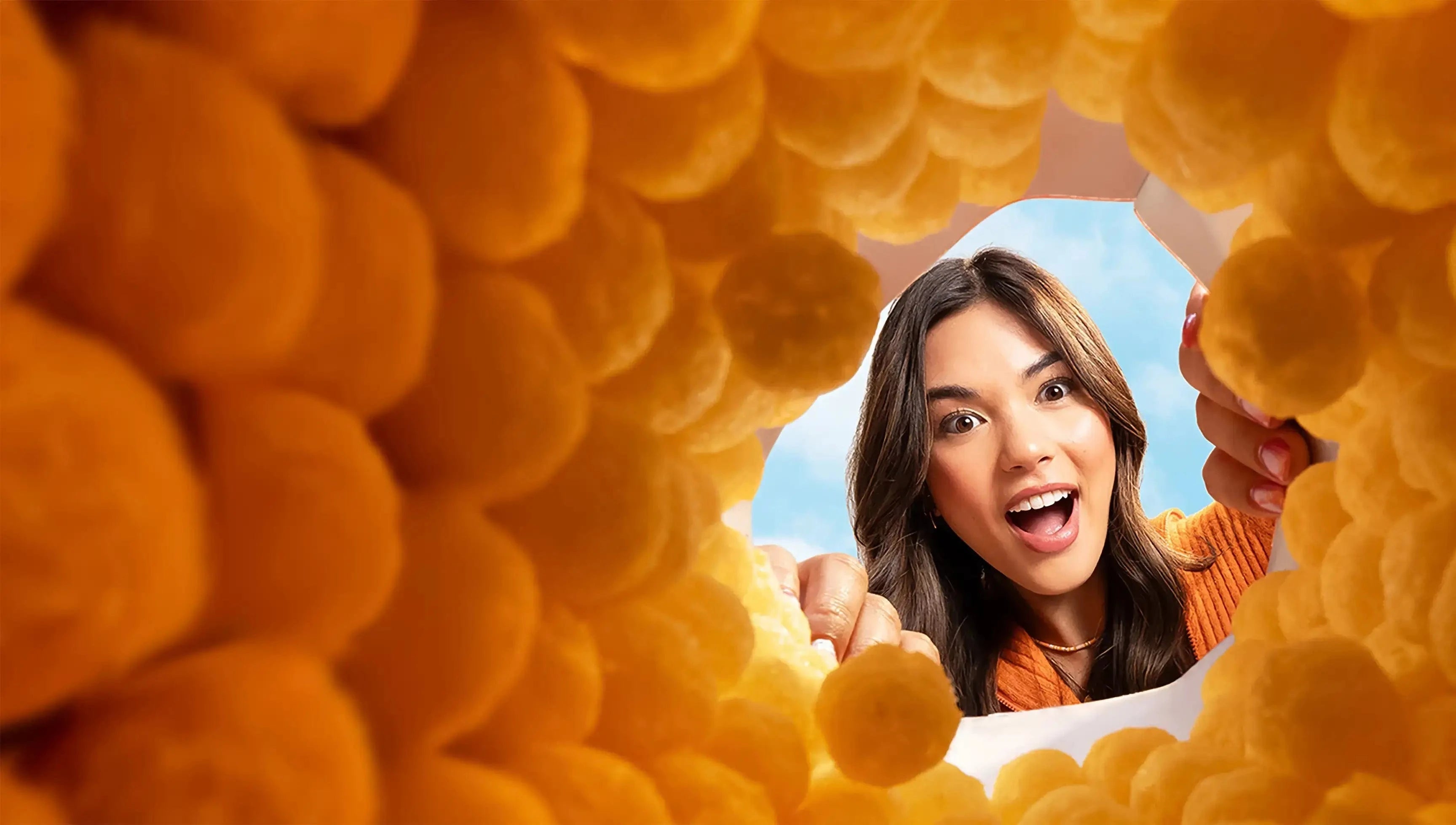 A surprised woman looks into a circular opening surrounded by yellow cheese puffs, with a blue sky in the background.
