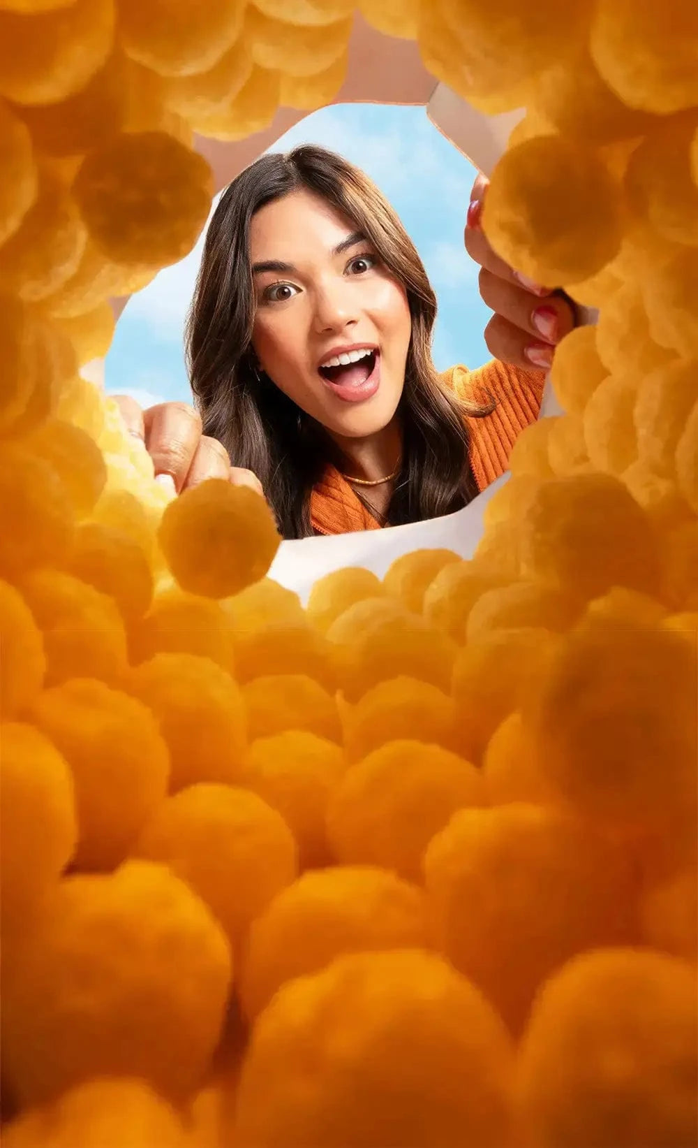 A woman smiles excitedly, reaching into a container filled with round cheese puffs, viewed from inside the container with the sky in the background.