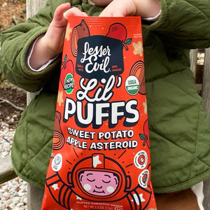 A child in a green jacket holds a bright orange bag of LesserEvil Sweet Potato Apple Asteroid Lil Puffs with space-themed cartoons and 0g added sugar on the packaging.