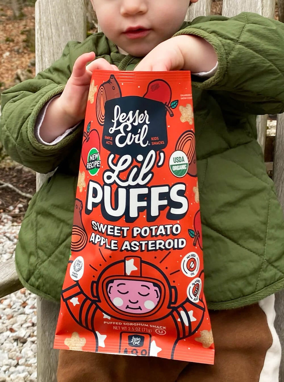A child in a green jacket holds a bright orange bag of LesserEvil Sweet Potato Apple Asteroid Lil Puffs with space-themed cartoons and 0g added sugar on the packaging.